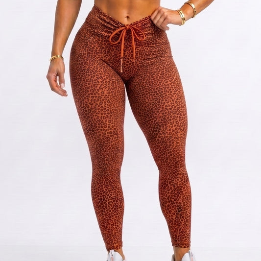 A woman modeling a matching burnt orange activewear set consisting of a halter-neck sports bra and high-waisted leopard-print leggings, paired with white sneakers, against a plain white studio background.