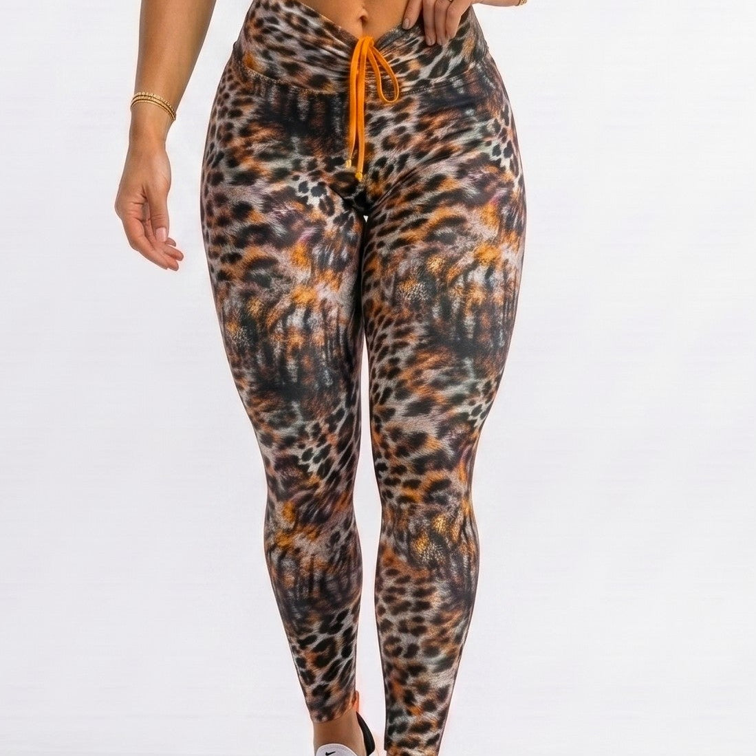 A woman modeling a matching activewear set consisting of a bright orange sports bra and high-waisted leopard-print leggings with an orange drawstring, paired with white sneakers, against a plain white studio background.