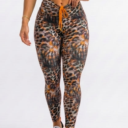 A woman modeling a matching activewear set consisting of a bright orange sports bra and high-waisted leopard-print leggings with an orange drawstring, paired with white sneakers, against a plain white studio background.